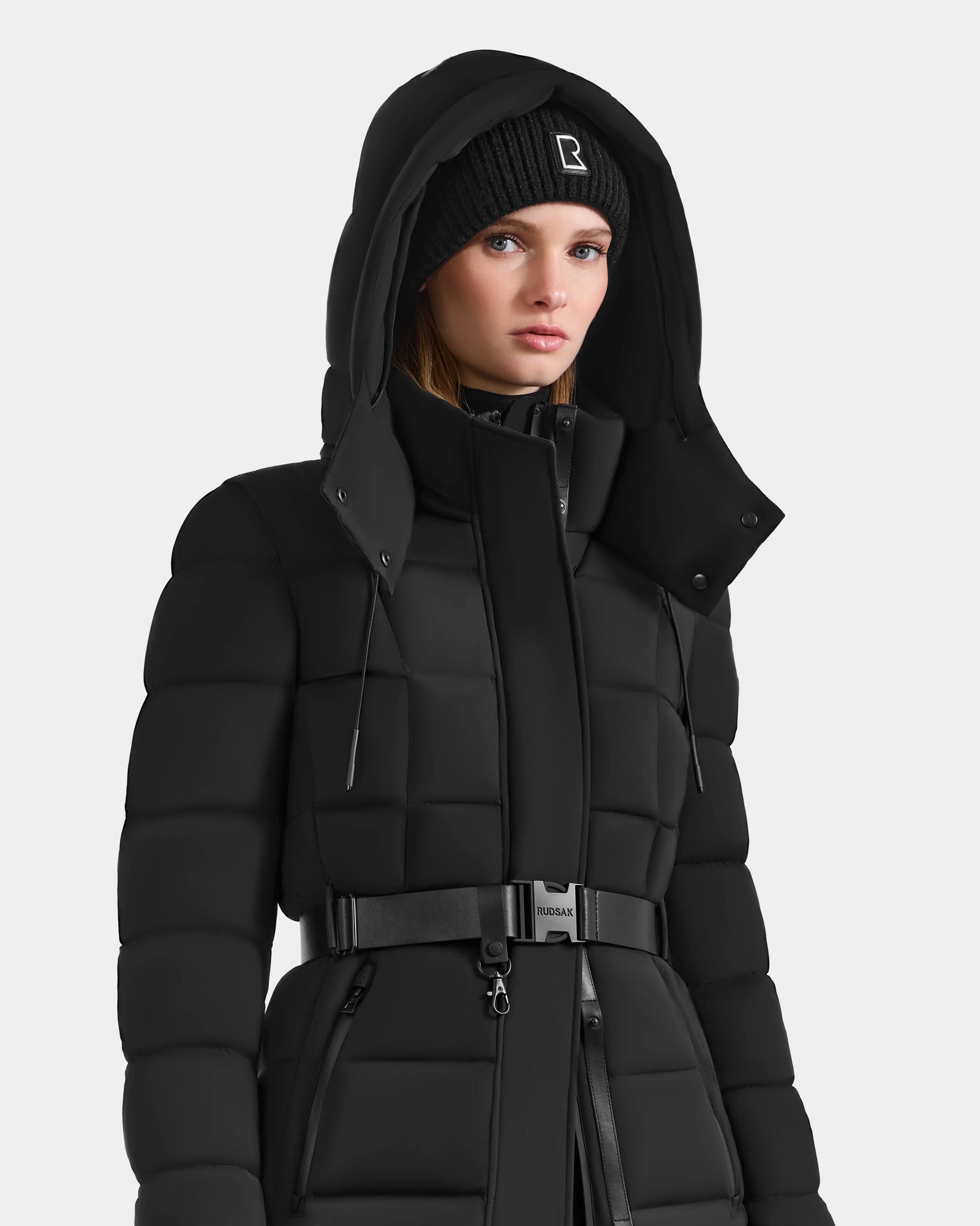 A woman in the Rudsak FRIDA M-M - L125607 Black quilted down puffer coat with detachable hood, belted waist, and a black knit logo beanie stands against a plain white background.