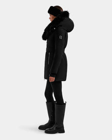 A person in profile models the Rudsak ISELLE - L125547 Black women’s down parka with a fur-trimmed hood, styled with black leggings, tall black boots, and a black headband against a plain light background.