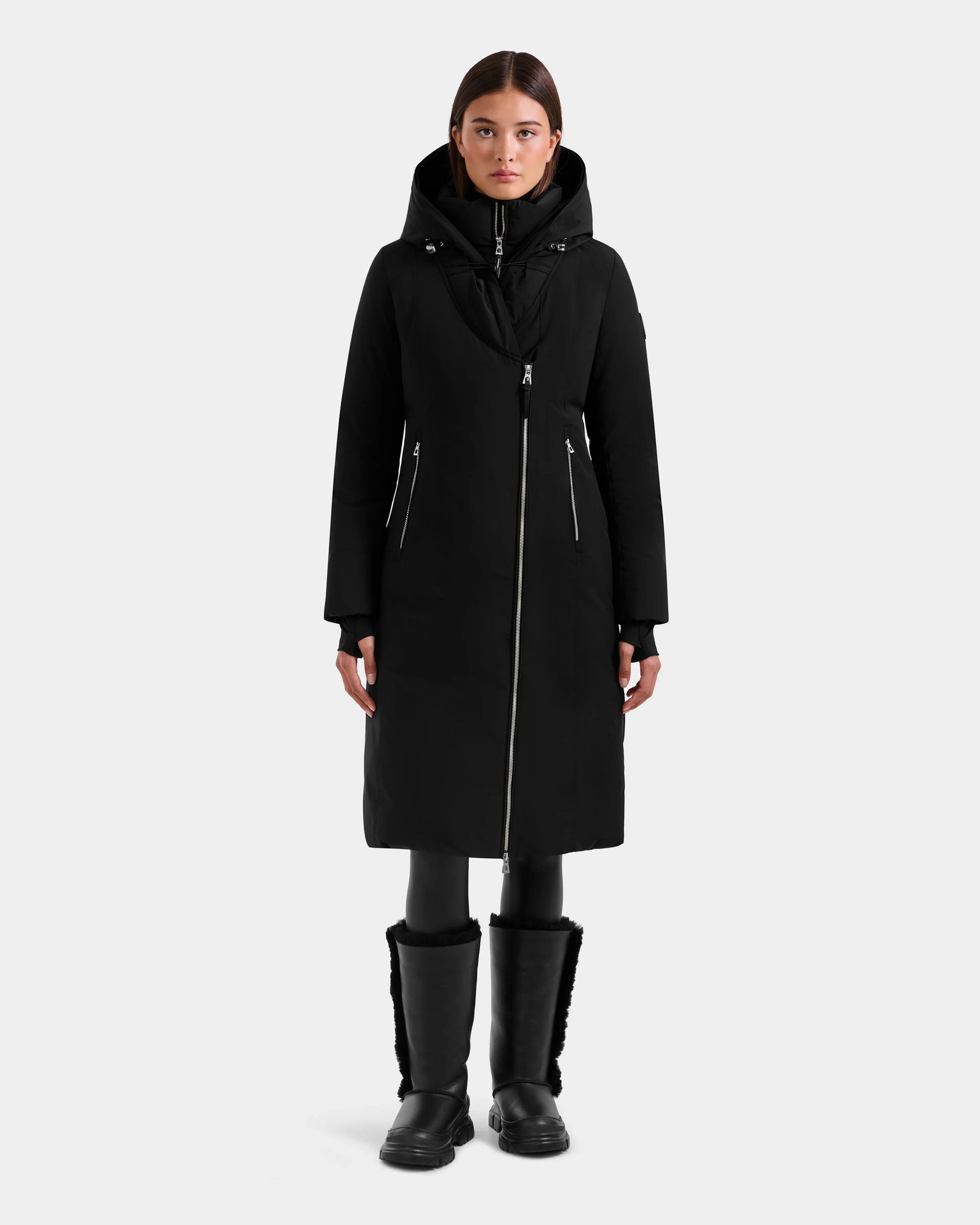 A woman faces forward against a plain white background, wearing the Rudsak JANIS - L125558 Black long winter coat with a faux fur hood, paired with black leggings and black boots.