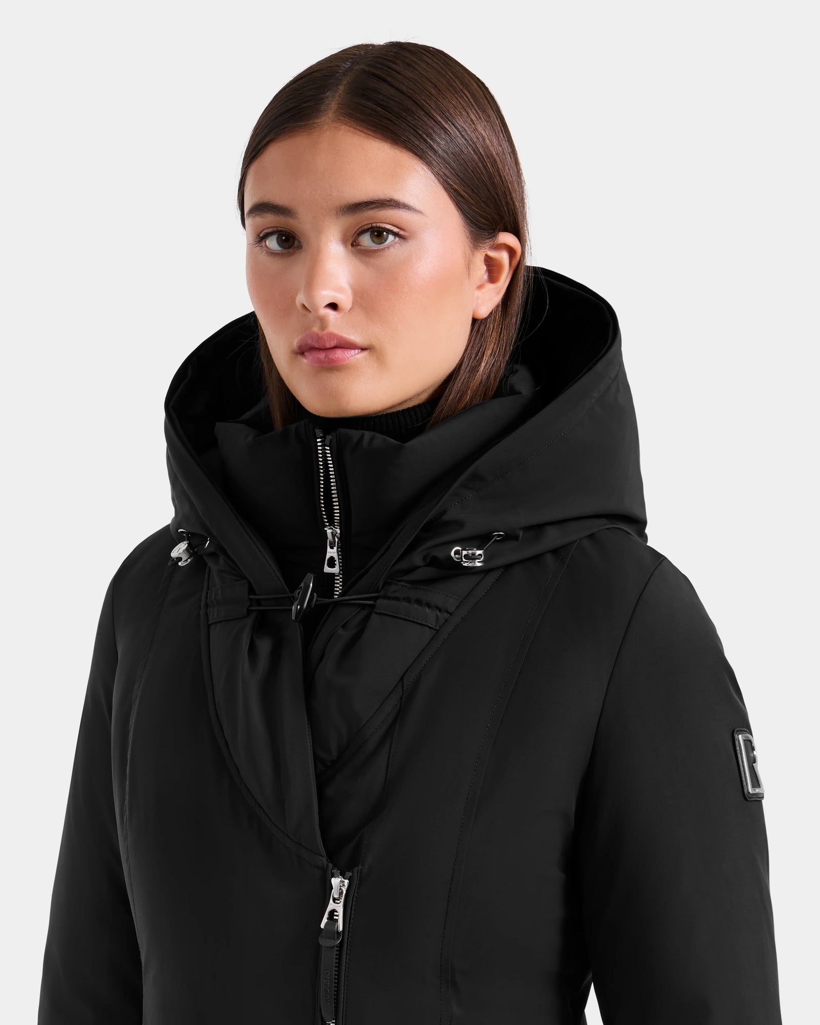 A young woman with straight brown hair models the Rudsak JANIS - L125558 Black long winter coat, featuring zippers and drawstrings, against a plain light gray background.