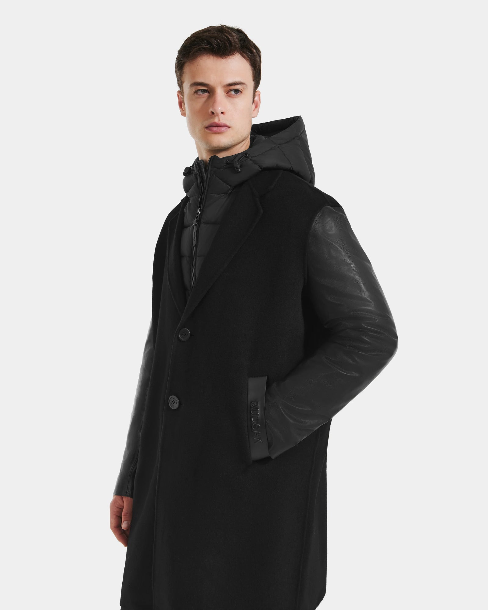 A man stands against a plain light background, wearing the Rudsak JESSE - M125504 Black men's wool coat with leather sleeves and a hooded inner jacket, one hand in his pocket and looking slightly to the side.