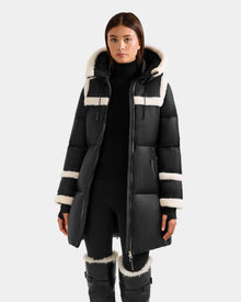 A woman models the Rudsak JULIANA L - L125535 Black/Vanilla puffer coat with white faux fur trim, belted waist, black pants, and boots—ideal for cold-weather style against a plain light background.