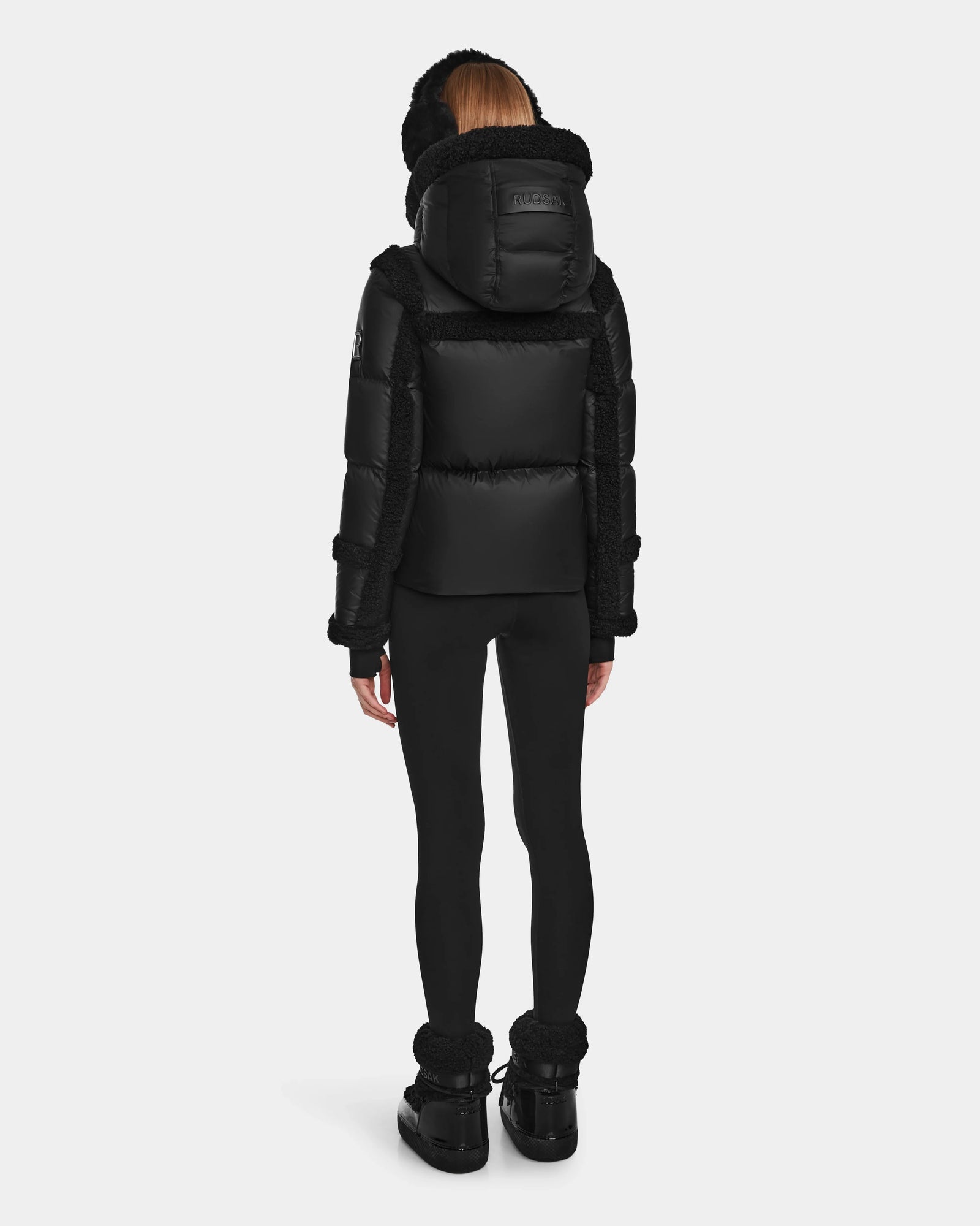 A person faces away, wearing the Rudsak JULIANA - L124509 Black/Black women’s short down puffer winter jacket with hood, black leggings, and black winter boots. RDS-certified down provides warmth. Set against a plain white background.