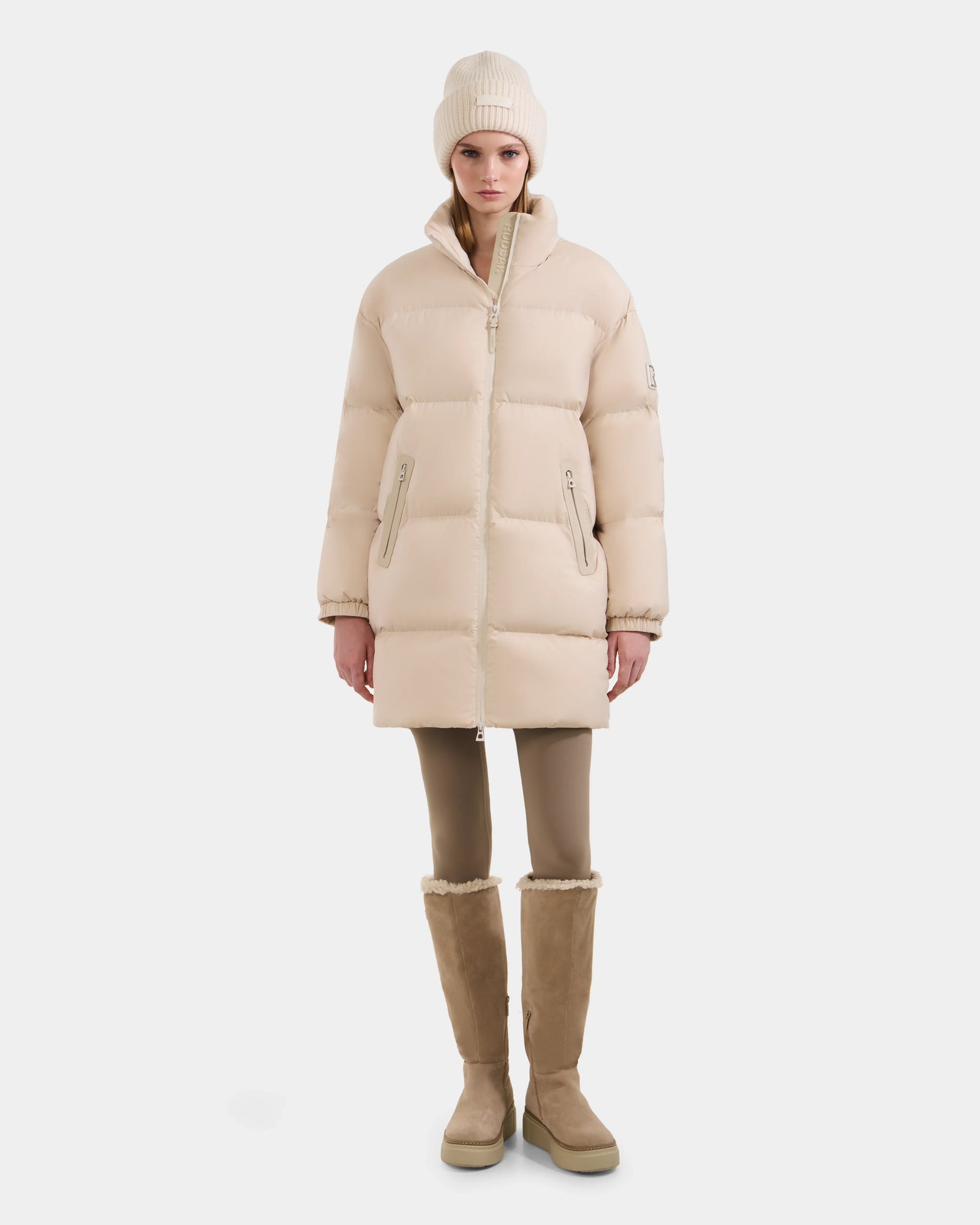 A woman stands facing forward against a plain white background, wearing the Rudsak KARLEE L - 8125682 Vanilla long down puffer with vegan leather trims, tan leggings, a beige knit beanie, and tan knee-high boots.