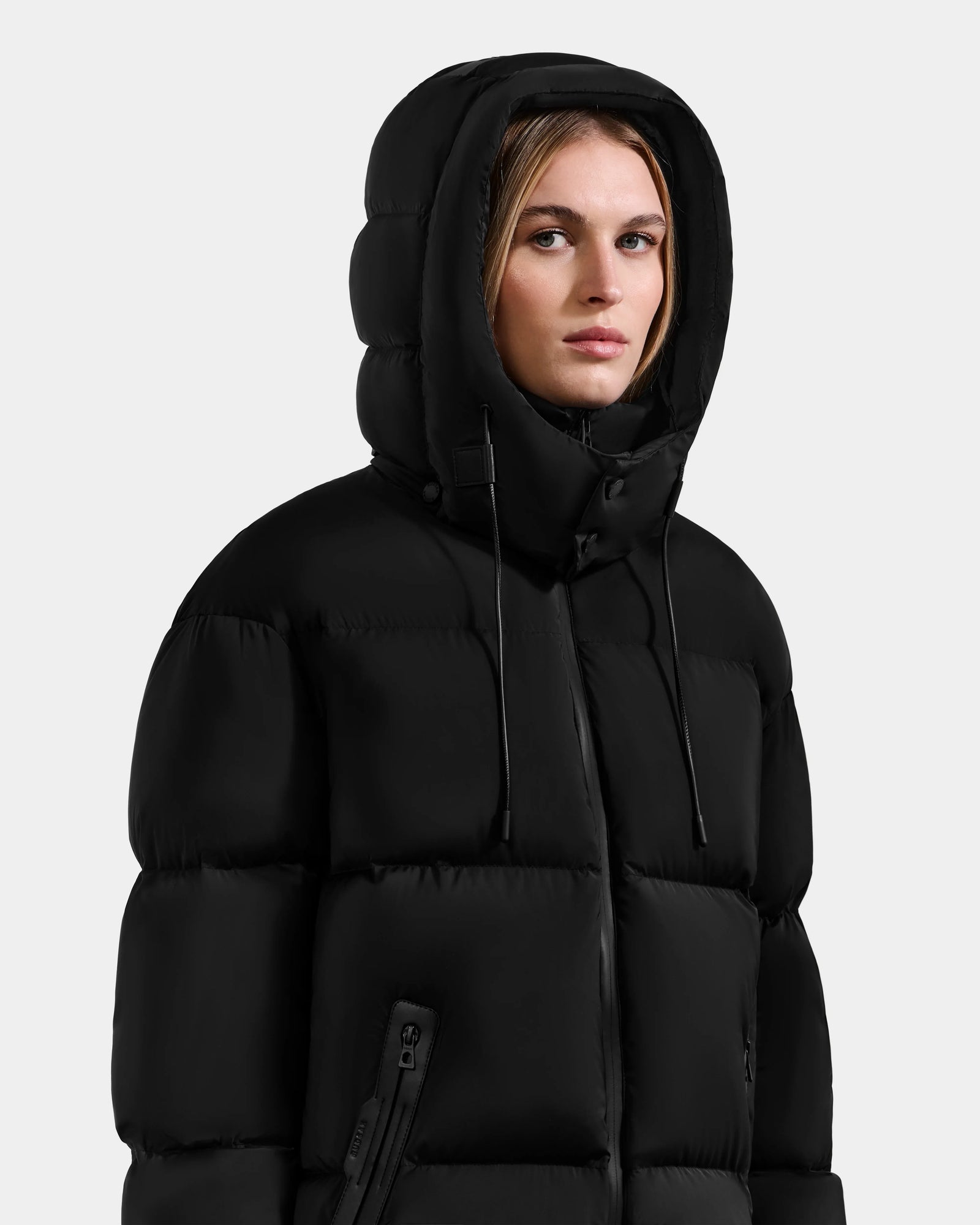 A person with straight, light brown hair and a neutral expression wears the Rudsak KARLEE M - 8125681 Black mid-length women’s down puffer jacket with a hood, quilted design, and zippered pockets, standing against a plain light gray background.