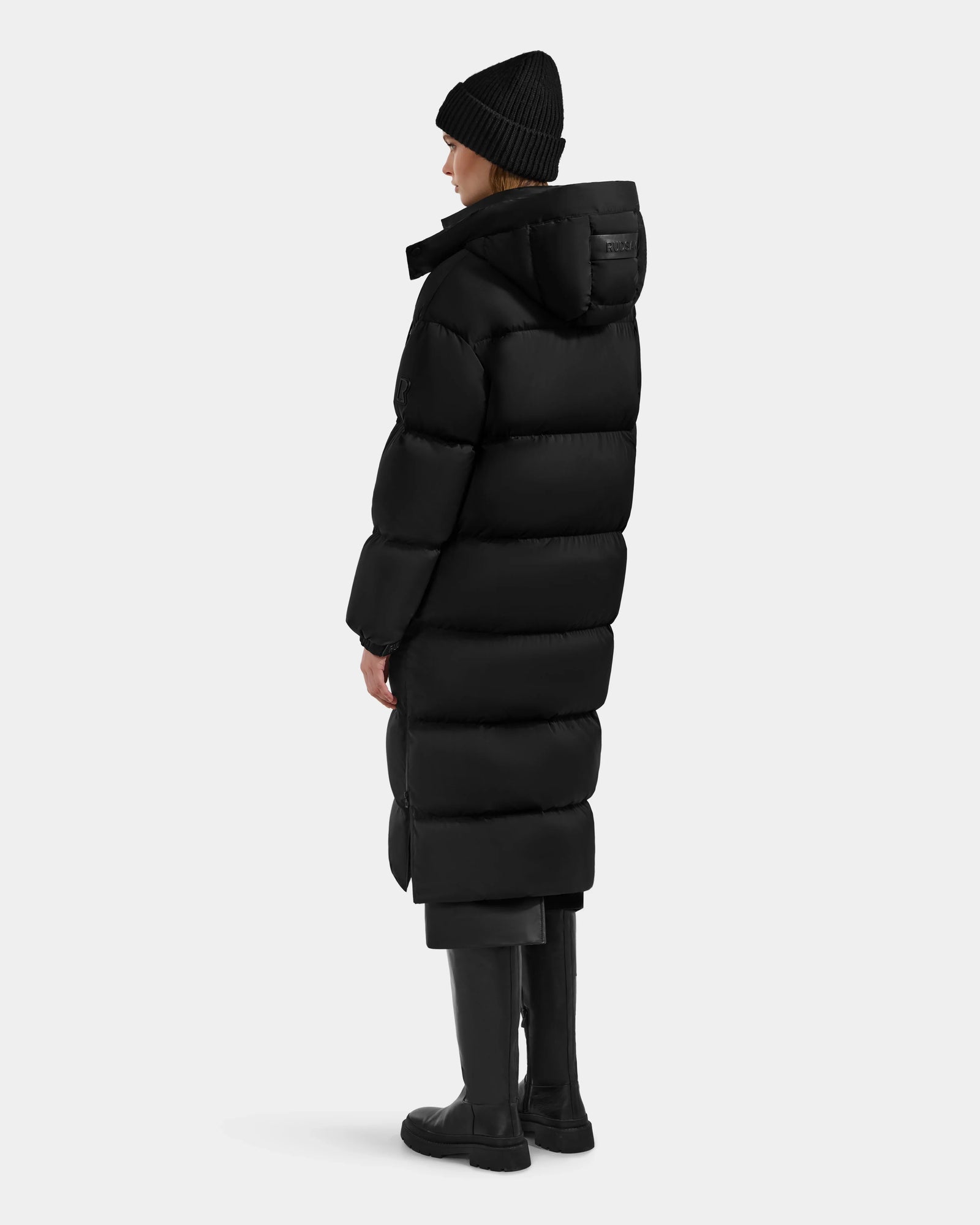 A person wearing the Rudsak KARLEE XL - 8125683 Black long maxi down puffer jacket stands facing away and slightly to the side against a plain light gray background—a true winter essential.