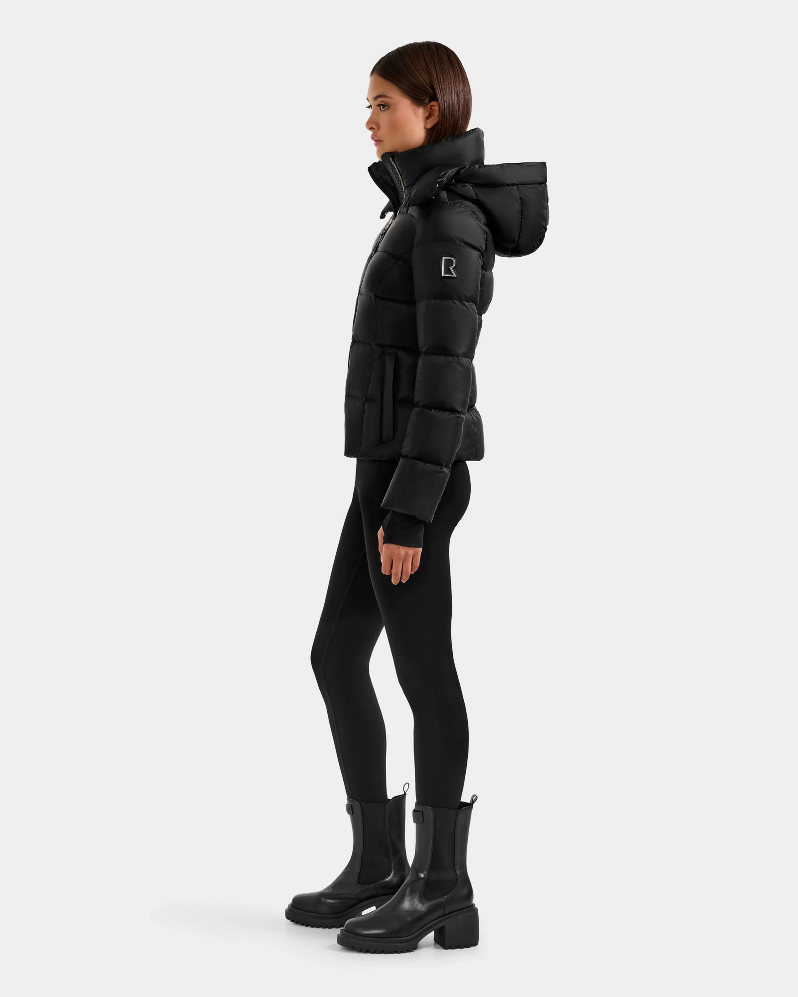 A woman stands in profile wearing the Rudsak KATIA - L125564 Black weather-resistant puffer jacket with a hood, paired with black leggings and chunky boots, set against a plain white background.