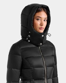 A woman in the Rudsak KATIA - L125564 Black puffer jacket with hood up, faces slightly sideways. The weather-resistant everyday jacket features a front zipper and buttoned hood. Background is light gray.
