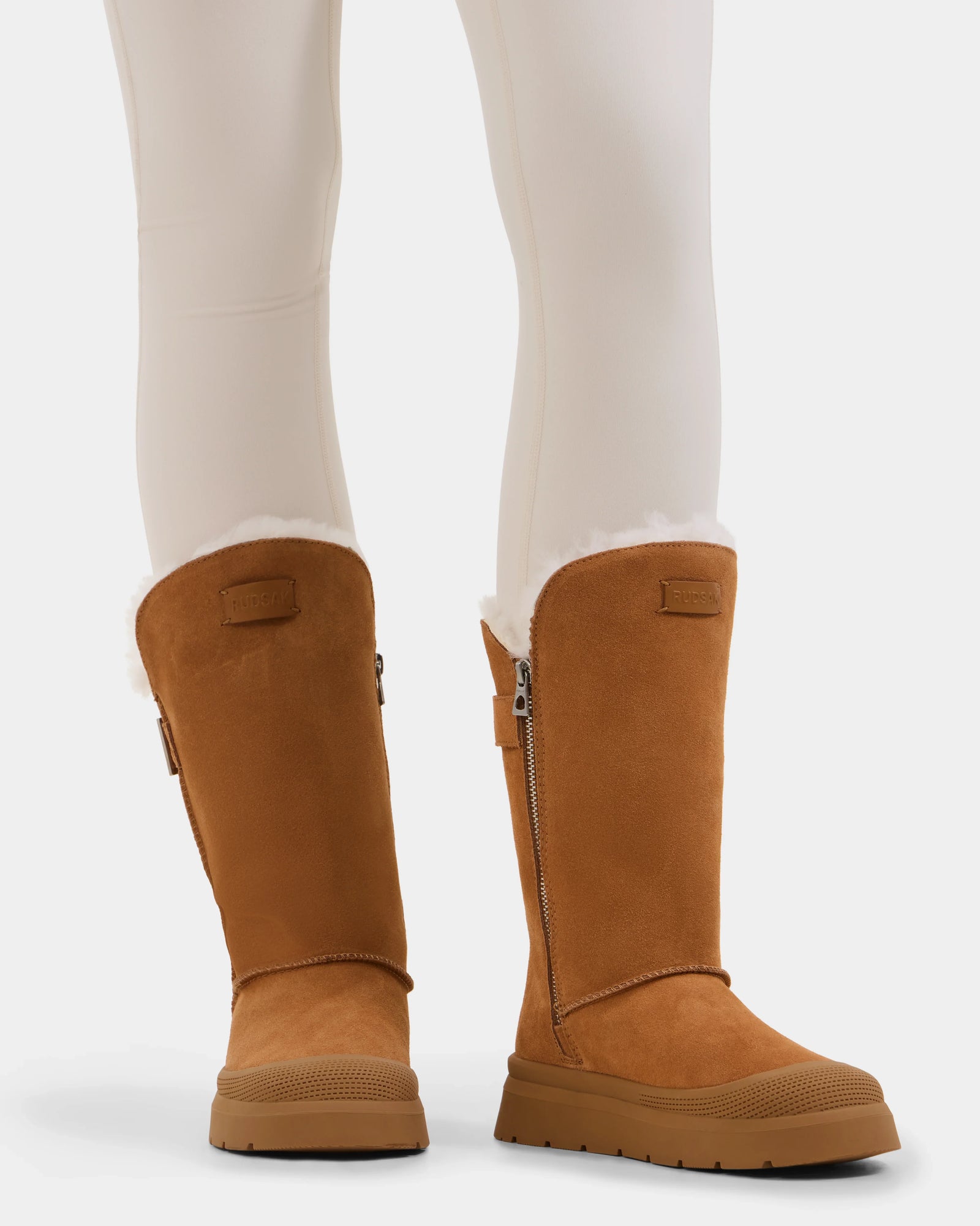 A person wears Rudsak KLAIR - L225027 Tan women’s mid boots—tall, tan suede winter boots with white wool lining, side zippers, and thick grooved rubber soles—paired with off-white leggings for extra warmth and style.