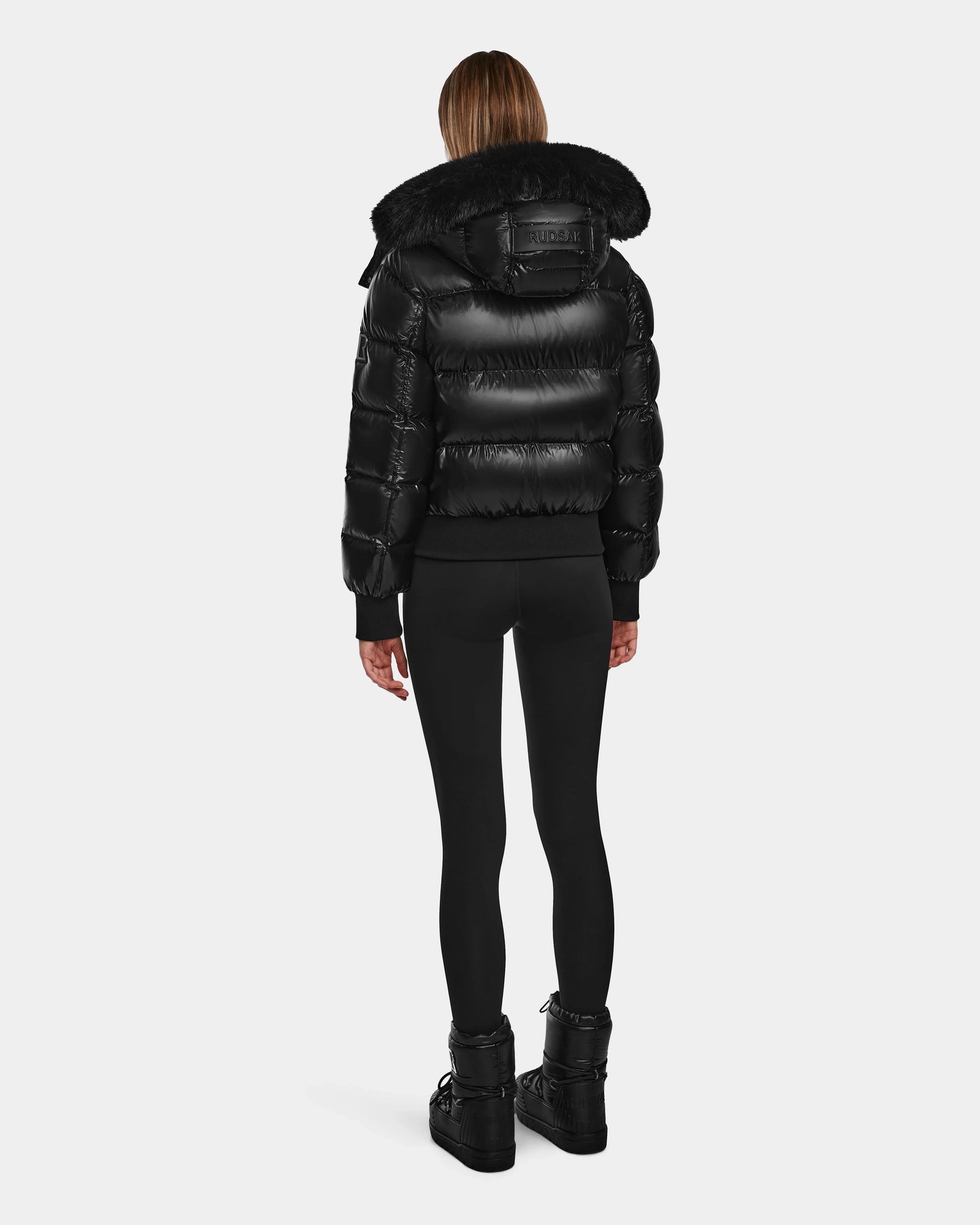 A person stands facing away, wearing the Rudsak MAUDE Q-S - L124553 Black shiny quilted down bomber with a fur-trimmed hood—this stylish winter jacket is perfect for cold days.
