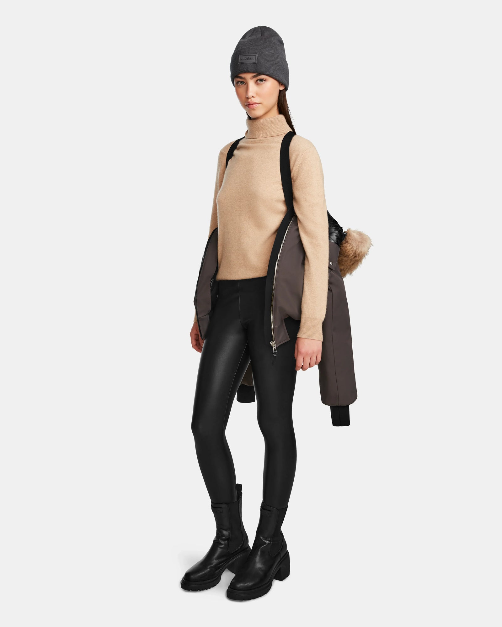 A woman wearing the MAUDE - L124522 Grey classic winter jacket from Rudsak drapes it over her shoulders, paired with a gray beanie, beige turtleneck, black leggings, and boots, standing with a neutral expression against a white background.
