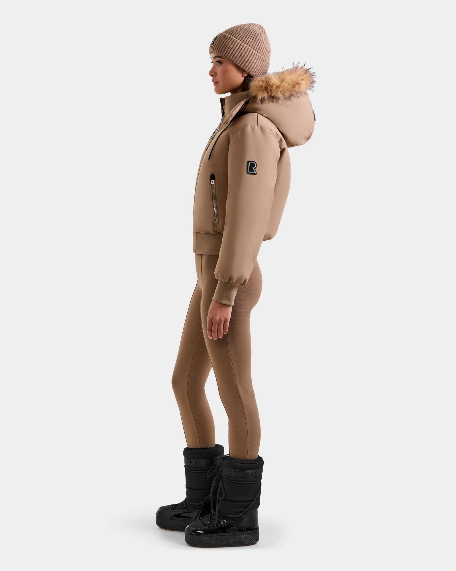 A woman in profile wears the Rudsak MAUDE - L124522 Sand down bomber, matching knit leggings, a tan beanie, and black winter boots, standing against a plain light background.