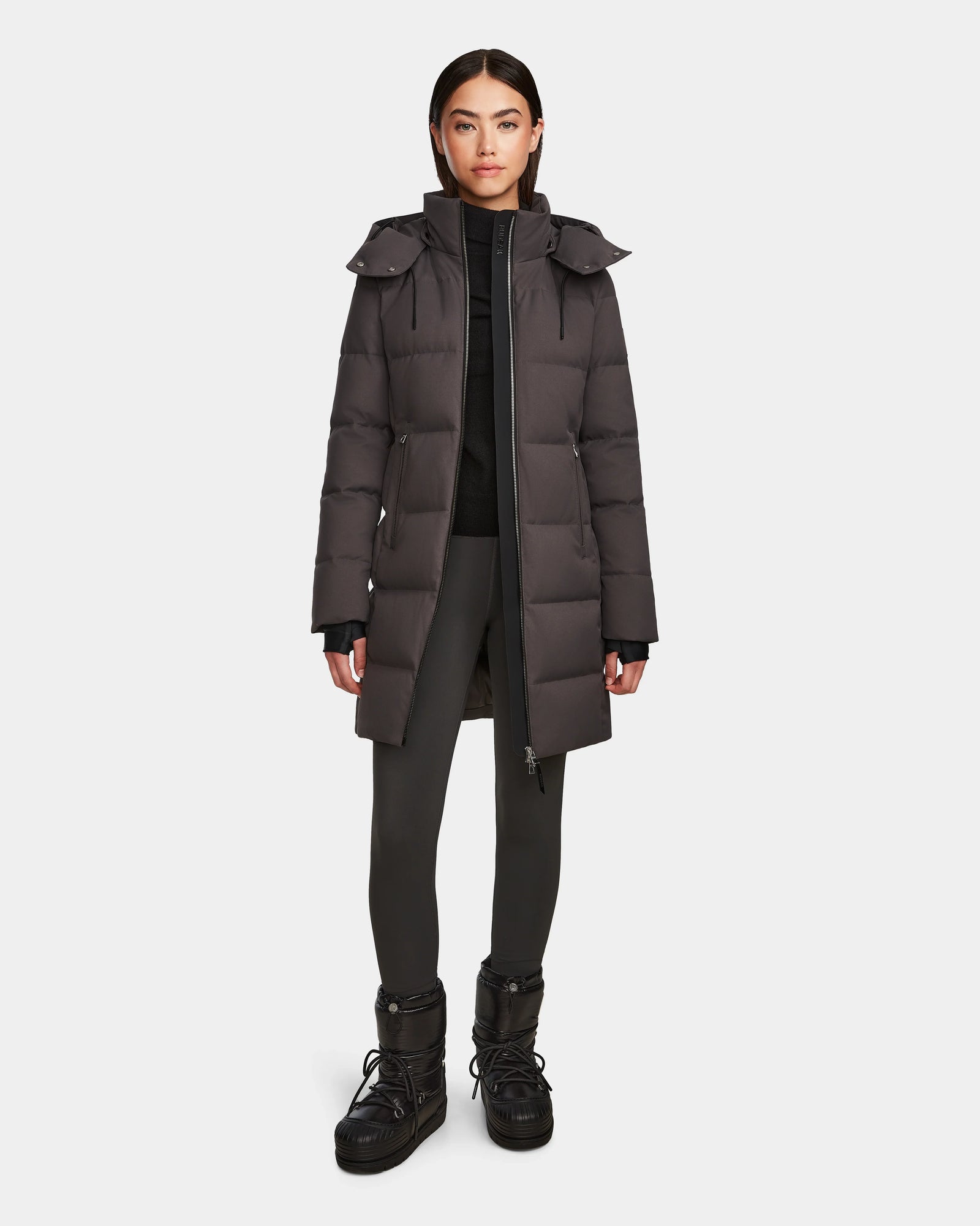 A woman stands facing forward, wearing the Rudsak MIRIAM - L123544 Grey women’s down puffer coat with a weather-resistant shell, paired with black pants and black winter boots against a plain white background.