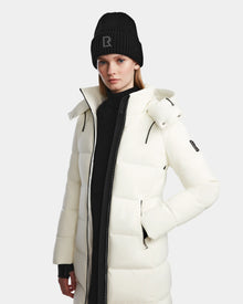 A woman wearing the Rudsak MIRIAM - L123544 Iceland white down puffer coat and a black beanie with an R stands against a plain light background, highlighting stylish winter essentials.