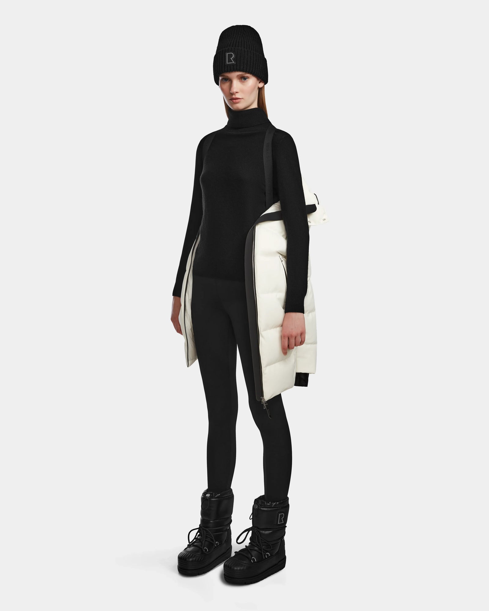 A woman stands against a plain background wearing a black turtleneck, leggings, beanie, and boots, with the Rudsak MIRIAM - L123544 Iceland women’s winter jacket draped over one shoulder.
