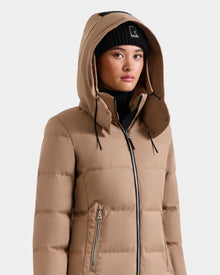 A person wearing the Rudsak MIRIAM - L123544 Sand women’s down puffer jacket with a hood and a black beanie looks slightly to the side. The weather-resistant jacket has zippered pockets and a high collar, set against a plain light gray background.