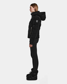A woman stands in profile wearing the MIRRY FF - L124507 Black women’s short down puffer jacket with a hood, black leggings, and black winter boots against a plain white background. The jacket features RDS-certified down for warmth and ethics.
