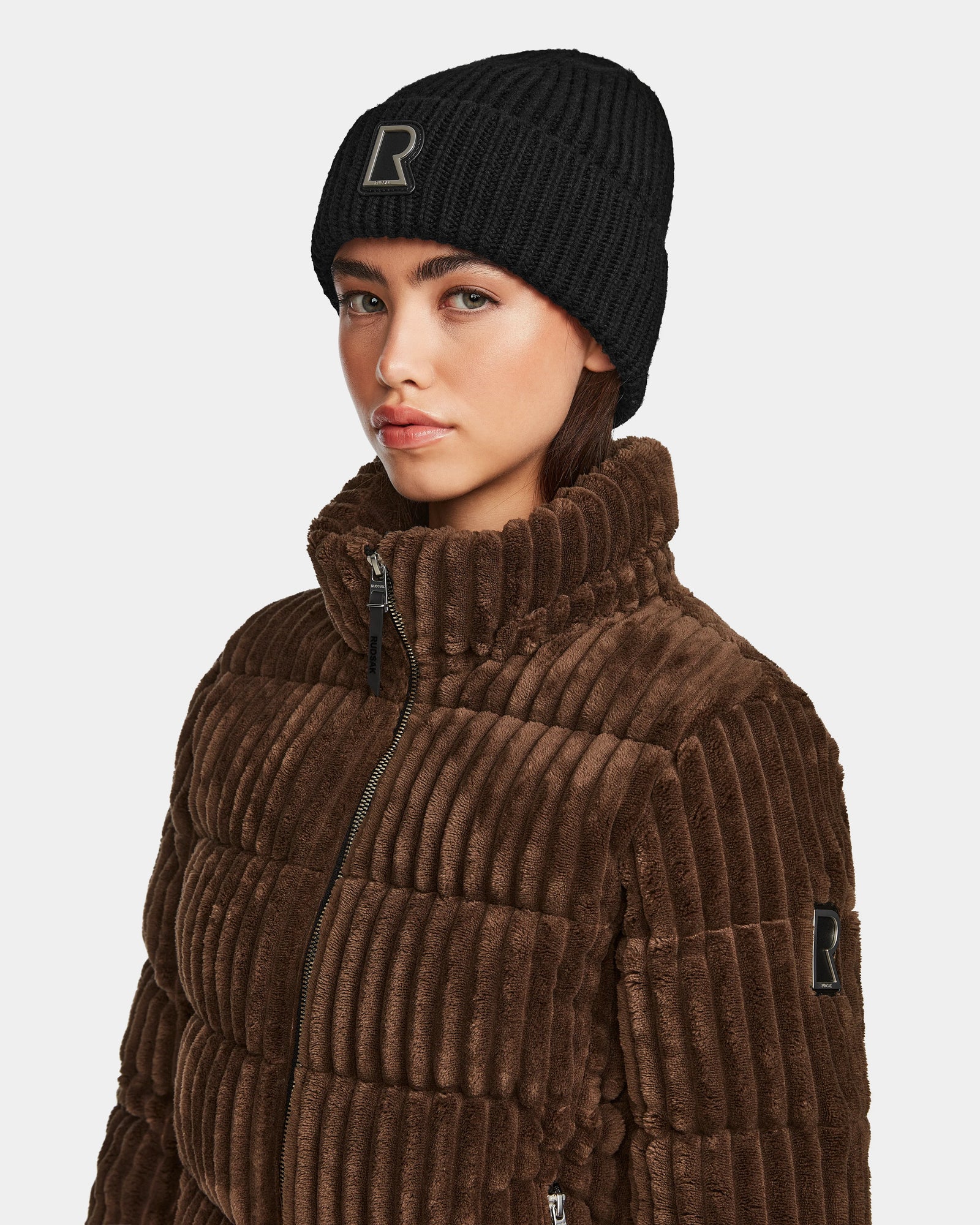 A woman wears a black knit beanie with an R patch and the Rudsak MIRRY FF - L124507 Espresso women’s short down puffer jacket with a high collar, standing against a plain light background.