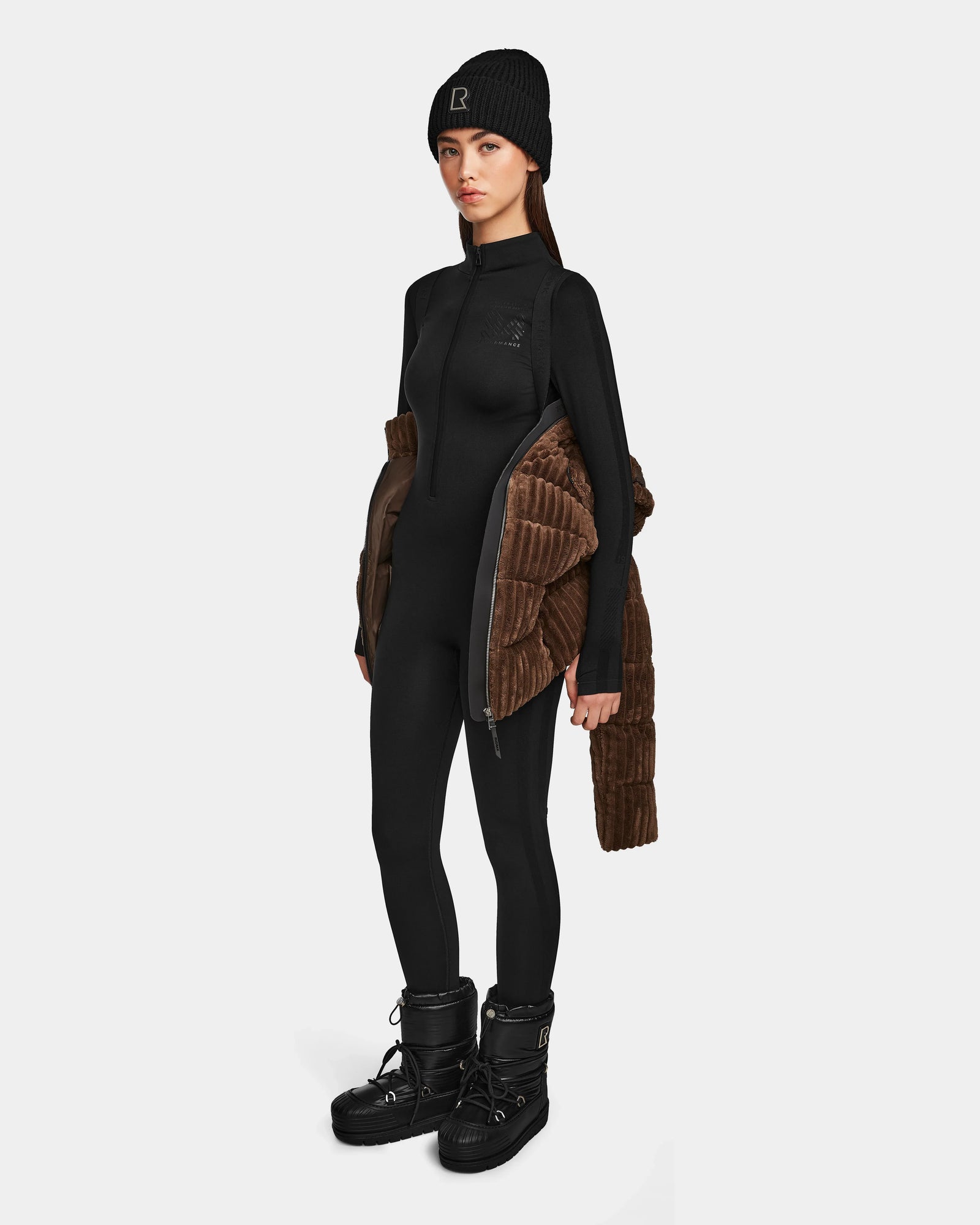 A woman stands against a white background wearing a black beanie, fitted black jumpsuit, black winter boots, and holding the Rudsak MIRRY FF - L124507 Espresso women’s short down puffer jacket draped over her arms.