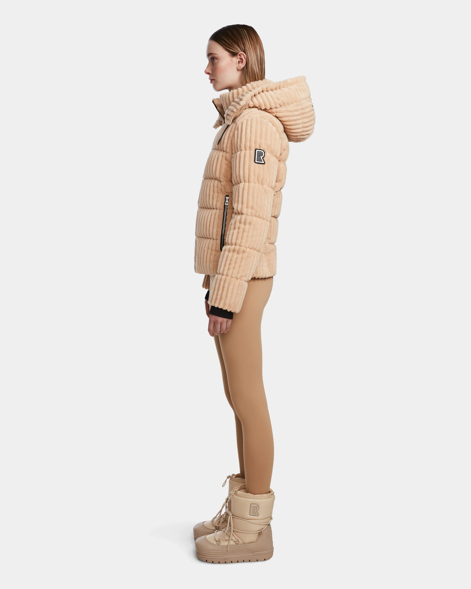 A person in profile wears the Rudsak MIRRY FF - L124507 Iceland beige velvet corduroy puffer jacket with a hood, patch sleeve, and vertical quilting, paired with tan leggings and beige boots against a plain white background.