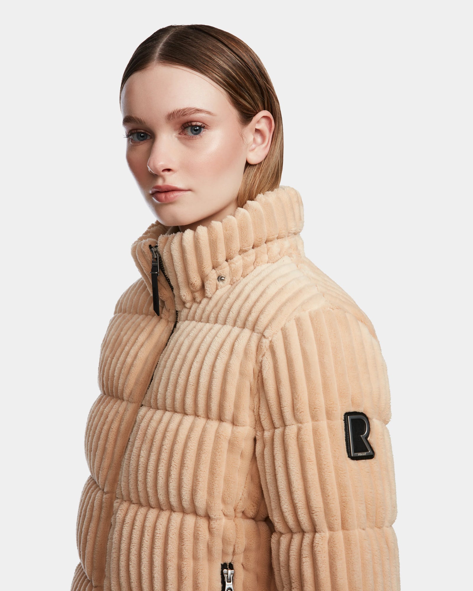 A woman with straight, light brown hair wears the Rudsak MIRRY FF - L124507 Iceland short down puffer jacket in beige with a ribbed finish, high collar, and black sleeve patch, posing against a plain white background.
