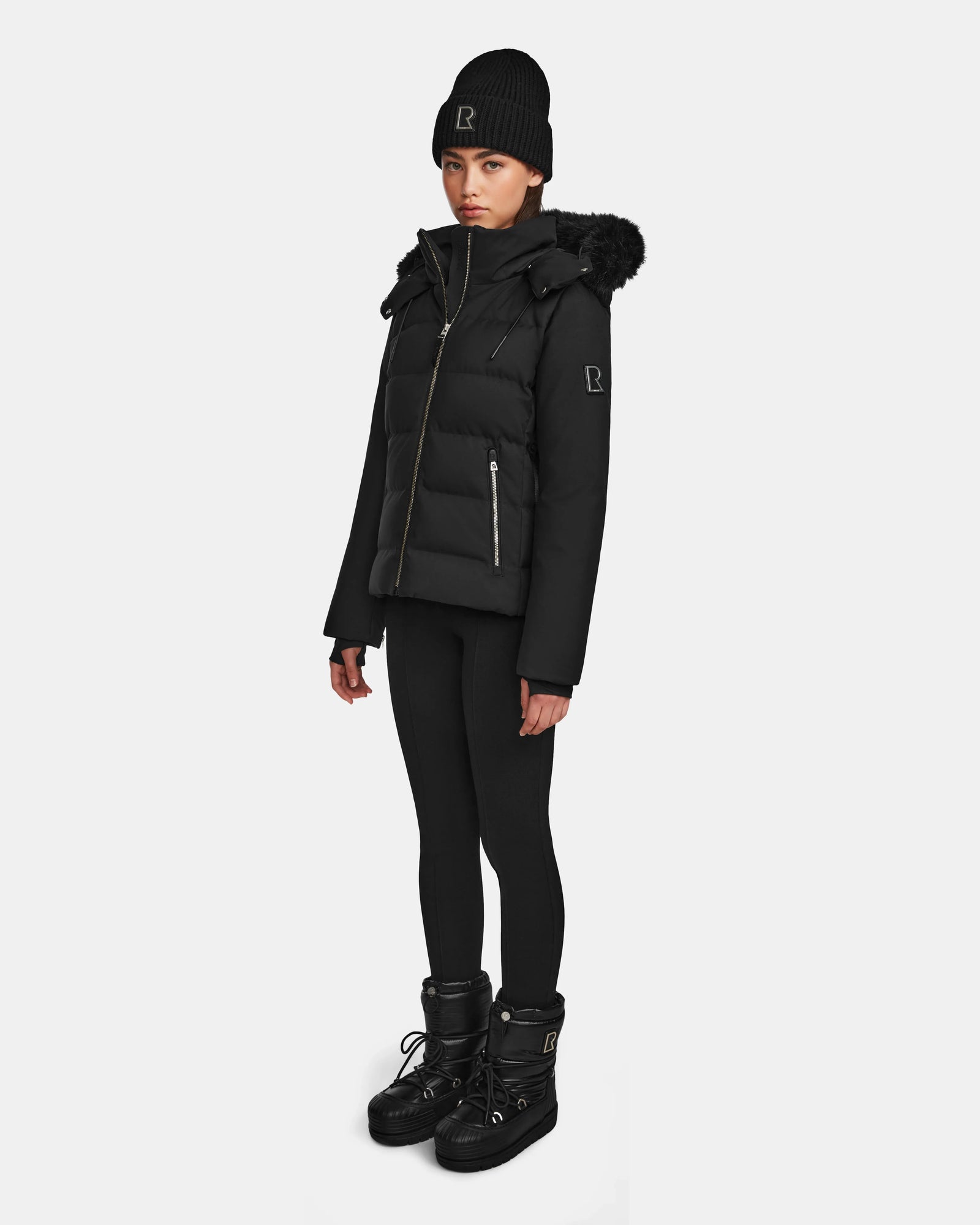 A woman wearing the Rudsak MIRRY M - L124533 Black quilted down coat with fur-trimmed hood, plus a black knit beanie, leggings, and winter boots stands against a plain white background.