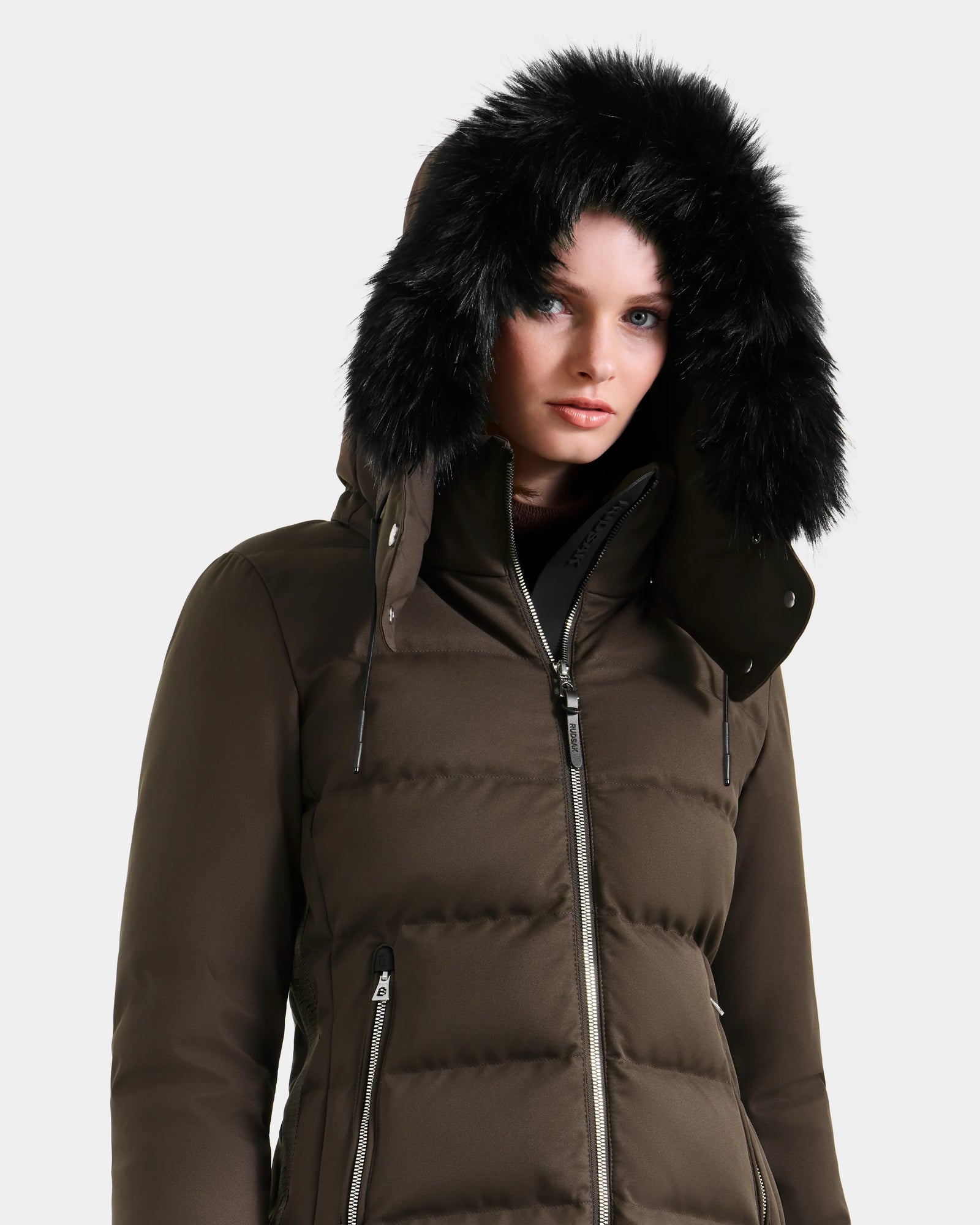 A person wearing the Rudsak MIRRY M - L124533 Espresso women's quilted down coat, a dark brown puffer jacket with a zip front and black faux fur hood, stands against a plain light background.