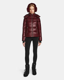 A woman faces forward in a shiny wine Rudsak MIRRY S - L123545 womens quilted down coat, with matching earmuffs, black leggings, and black winter boots, standing against a plain white background.