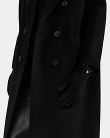 A person wears the Rudsak OLGA - L425541 Black long, double-breasted coat with women's wool gloves, one hand tucked in a pocket. The plain white background highlights this cold-weather essential look.