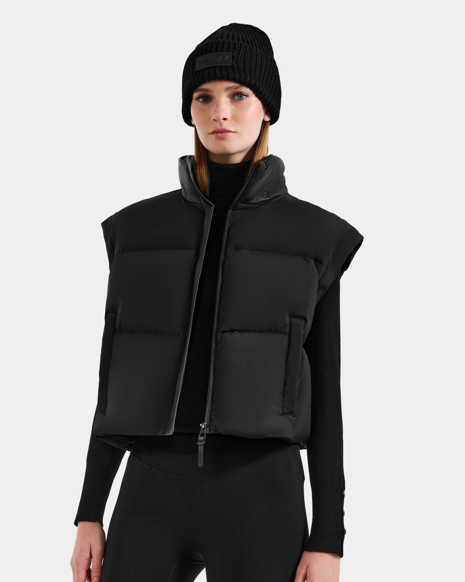 A woman in the Rudsak OPRAH - L125546 Black women’s cropped down puffer with removable sleeves and a black beanie stands against a plain light gray background, looking straight ahead with a neutral expression.