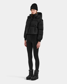 A person wearing the Rudsak OPRAH - L125546 Black women’s cropped down puffer with removable sleeves, black beanie, leggings, and winter boots stands against a plain light background, looking straight ahead with a neutral expression.