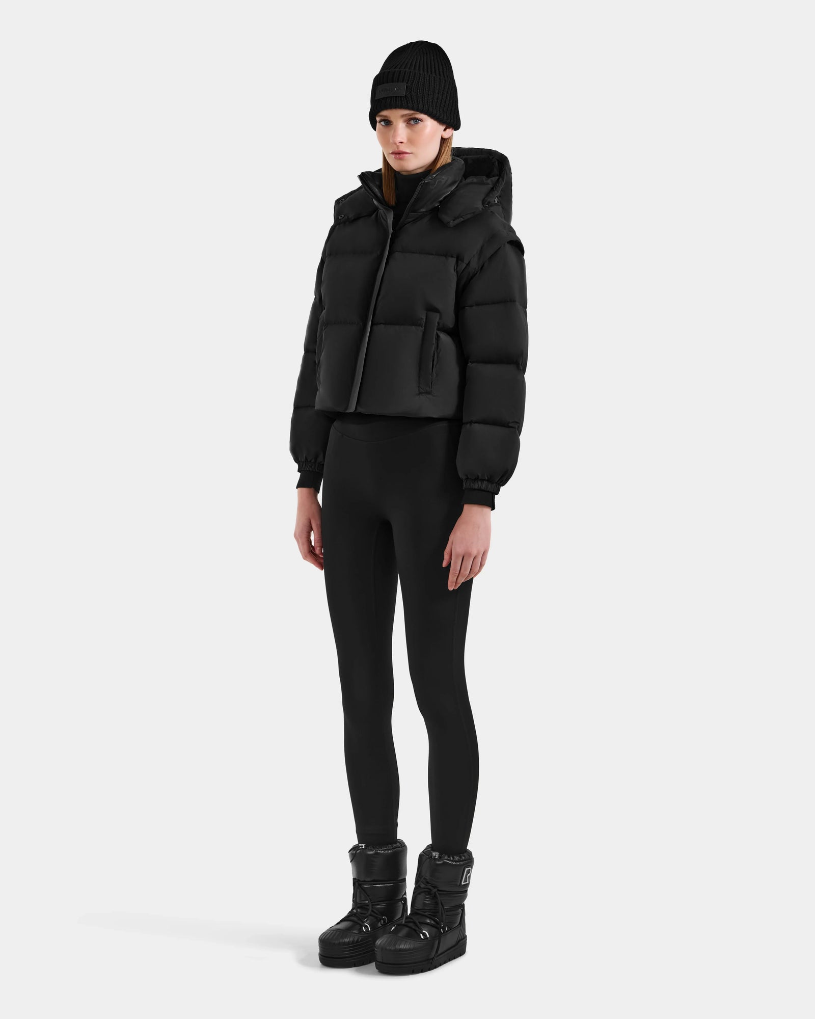 A person wearing the Rudsak OPRAH - L125546 Black women’s cropped down puffer with removable sleeves, black beanie, leggings, and winter boots stands against a plain light background, looking straight ahead with a neutral expression.