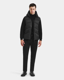 A man faces forward in the Rudsak RICHARDSON - M125527 Black men’s hooded vest over a black turtleneck, black pants, and shoes. Its lightweight warmth and water-repellent finish stand out against a plain light background.
