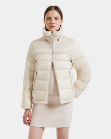 A woman with straight brown hair stands with one hand in her pocket, wearing the Rudsak SANDRA - 8125612 Vanilla shearling puffer jacket. She styles its high collar with a matching skirt and top for a modern, coordinated look.