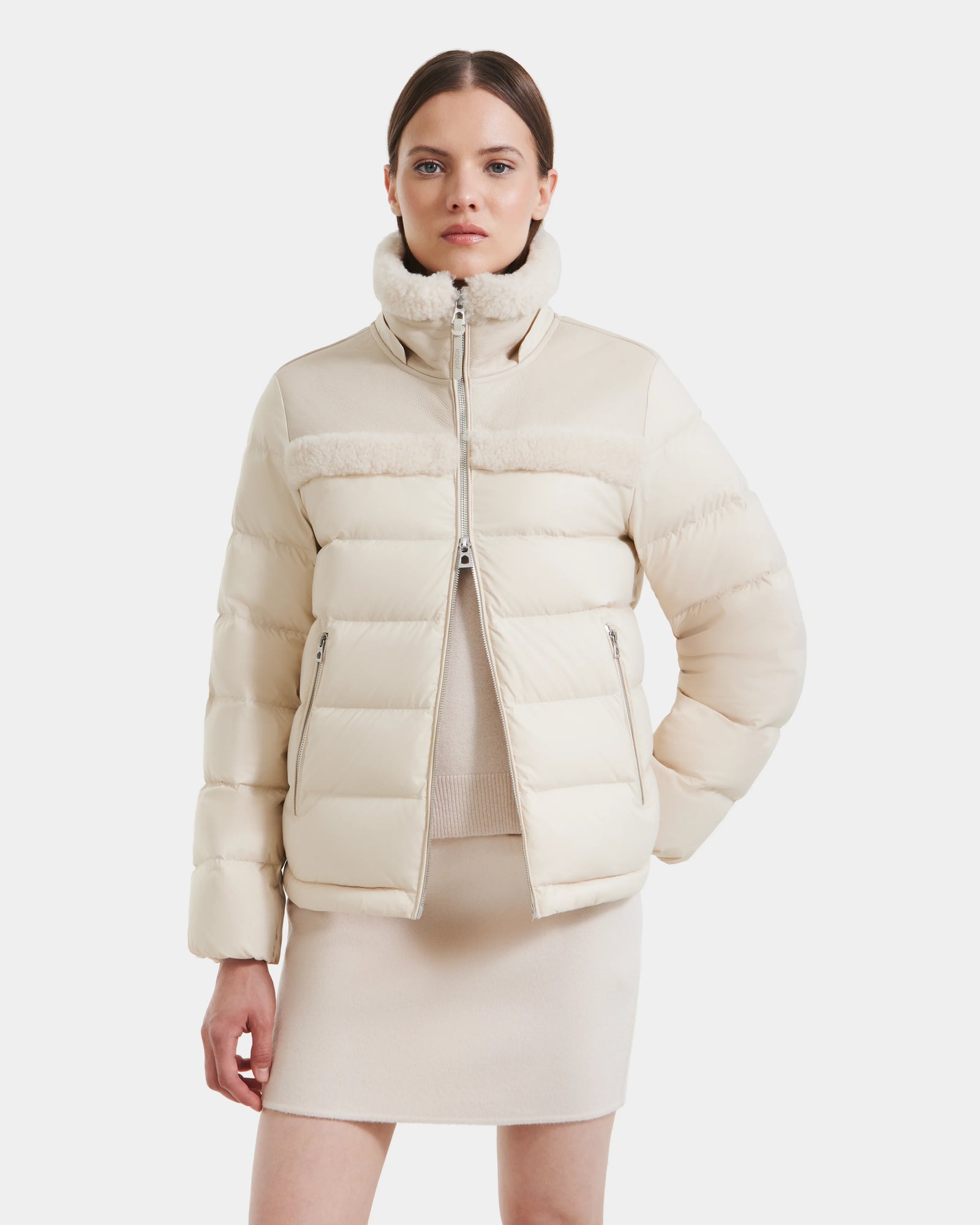 A woman with straight brown hair stands with one hand in her pocket, wearing the Rudsak SANDRA - 8125612 Vanilla shearling puffer jacket. She styles its high collar with a matching skirt and top for a modern, coordinated look.