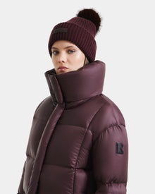 A woman wearing the Rudsak SASHA - 8125686 Bordeaux down puffer jacket with a high collar and a matching pom-pom knit beanie stands against a plain light background.