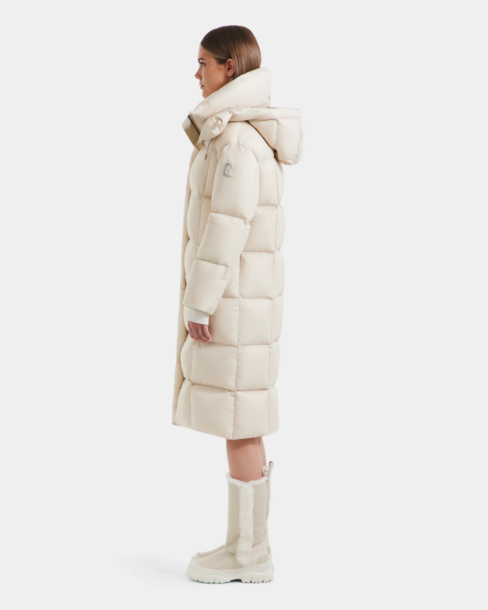 A woman stands in profile wearing the Rudsak SASHA - 8125686 Vanilla long down puffer coat with a large, removable hood and matching knee-high winter boots against a plain white background.