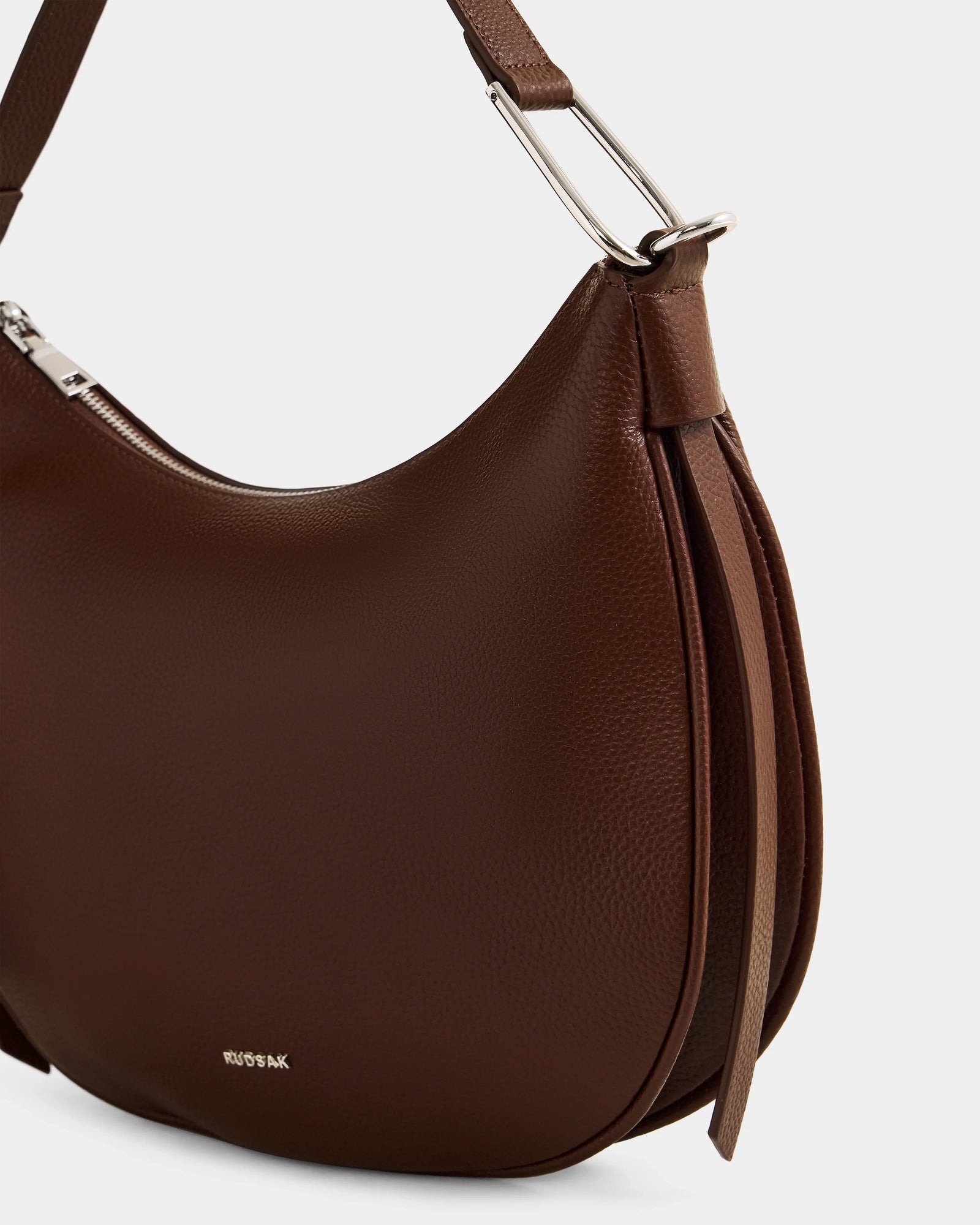 A close-up of the Rudsak SYLVIA - L325515 Mocha women’s leather shoulder bag shows its crescent shape, silver hardware, textured finish, small logo near the bottom, and two leather straps hanging from the side.