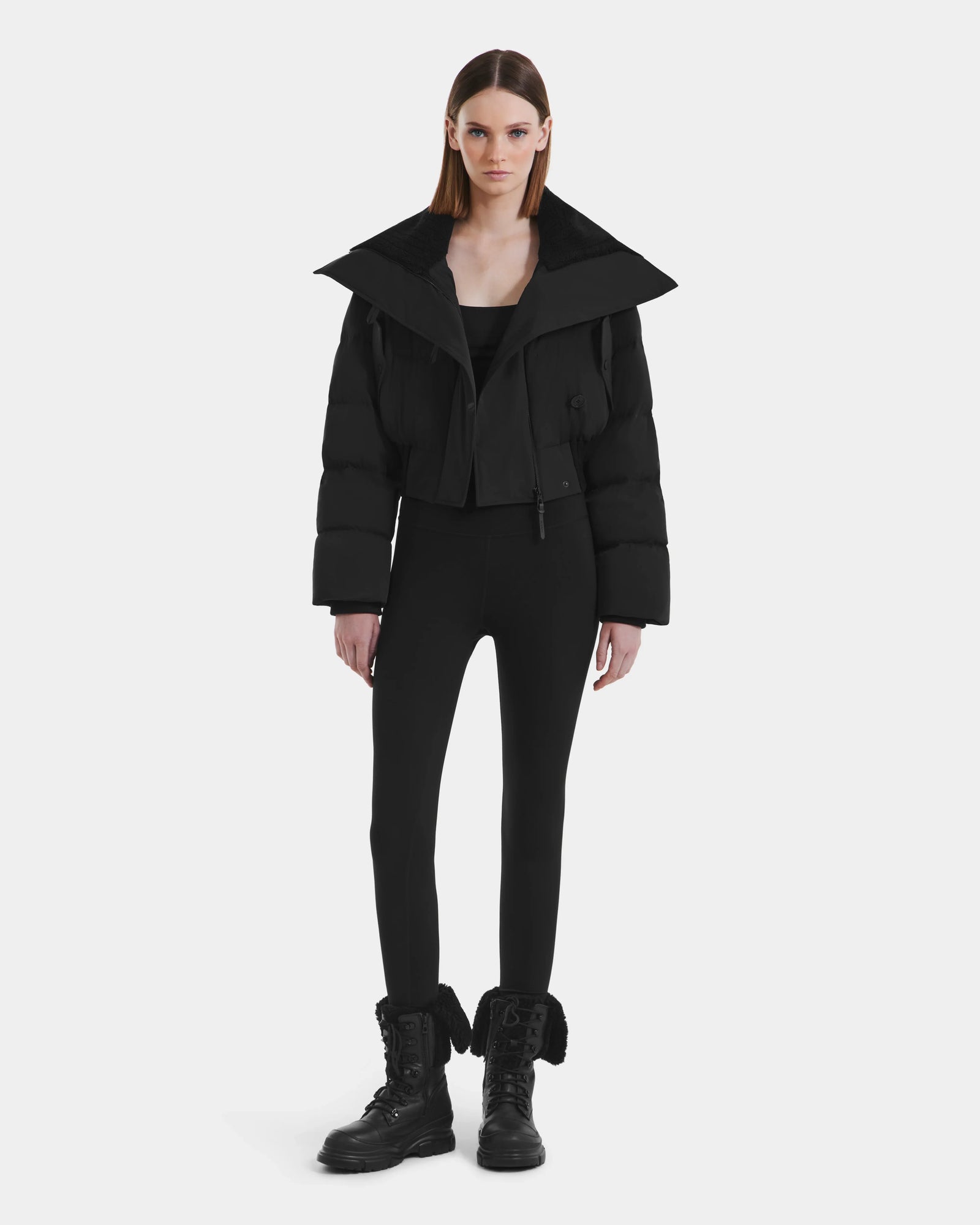 A woman faces forward against a white background, wearing the Rudsak YVETTE - L125557 Black cropped down jacket with an oversized collar, paired with black leggings and black combat boots. Her straight hair is down and her expression is neutral.