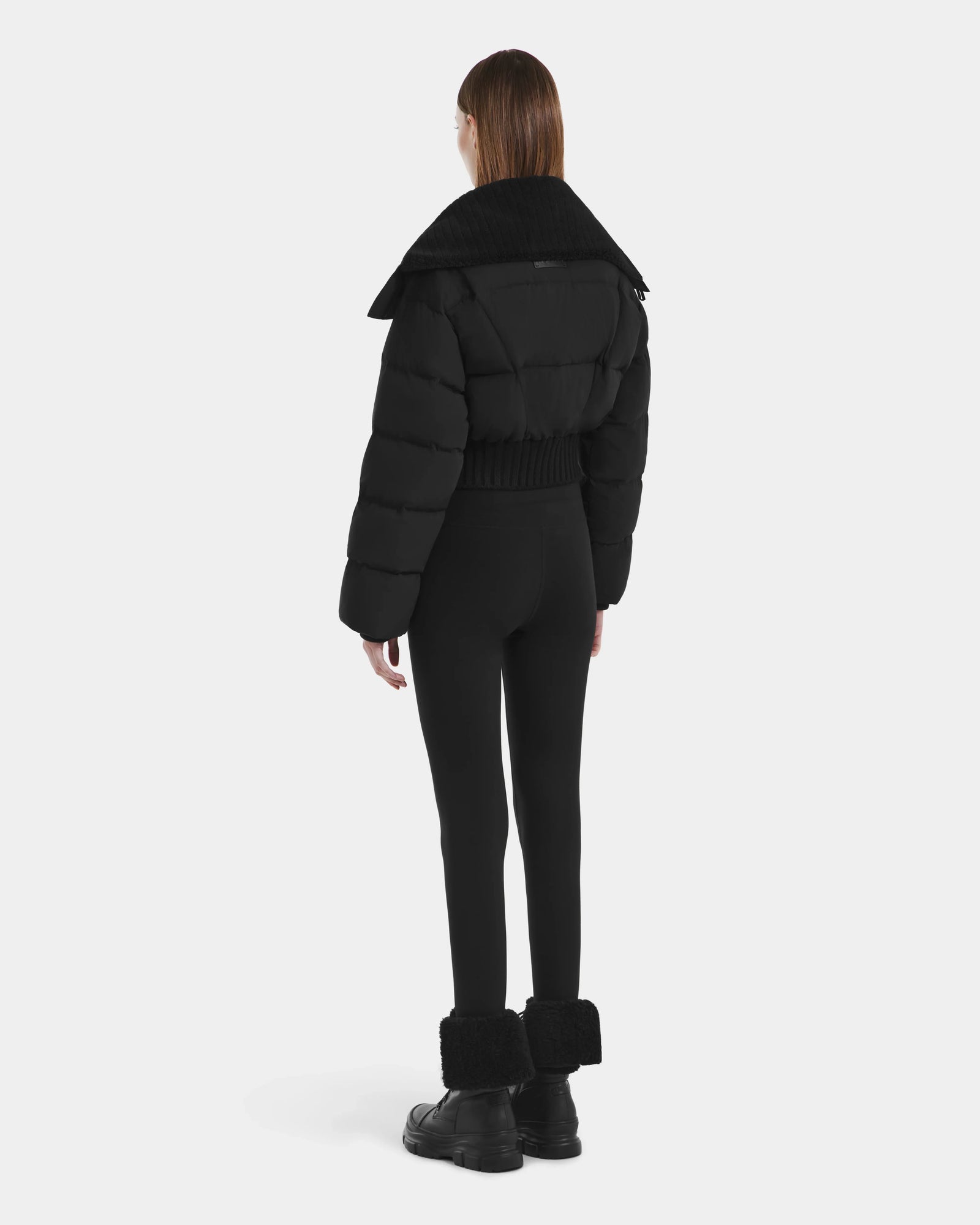 A person with straight brown hair, seen from behind, wears the Rudsak YVETTE - L125557 Black women’s cropped down jacket with an oversized collar, black leggings, and black fur-cuff boots against a plain white background.