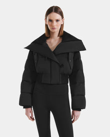 A woman poses against a white background wearing the Rudsak YVETTE - L125557 Black cropped down jacket with an oversized collar and fitted black high-waisted pants, looking directly at the camera.