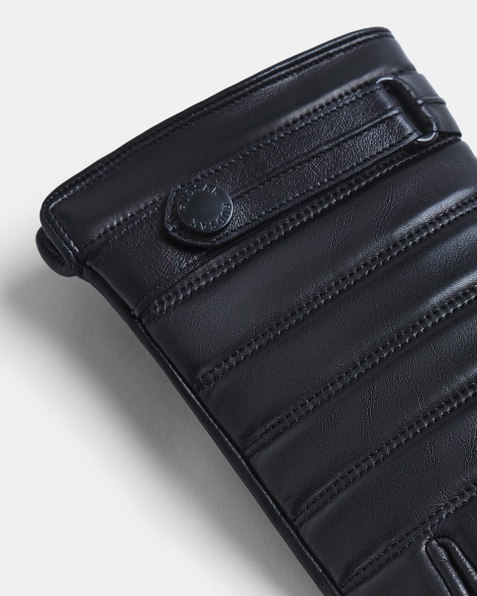 Close-up of the CORTES - 6418506 BLACK by Rudsak: black leather with horizontal padding, precise stitching, a snap button embossed with the Rudsak logo, smooth texture, and rounded edges—reflecting premium craftsmanship in Rudsak gloves.