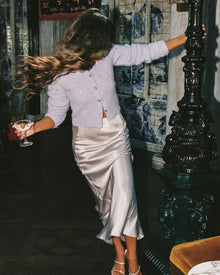 Lifestyle back view image of a model wearing the LSPACE Sweet As Honey Skirt in silver, a fluid satin midi skirt with a bias cut, styled with a pale blue embellished cardigan while holding a martini glass in an ornate bar setting with blue-and-white tiled walls.