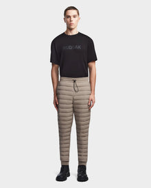 A young man stands facing forward against a white background, wearing Rudsak’s MIKE - 6523503 Cement men’s beige quilted drawstring pants in stretch neoprene, a black Rudsak t-shirt, and black shoes.