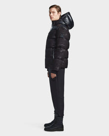 A young person stands in profile wearing the Rudsak RONNY M - M123512 Camo Black hooded down puffer jacket with black pants and boots, against a plain light gray background.