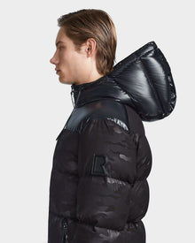 A young man with light brown hair, wearing the Rudsak RONNY M - M123512 Camo Black weather-resistant hooded down puffer jacket with a shiny hood and raised R patch on the sleeve, is shown in profile against a plain light background.