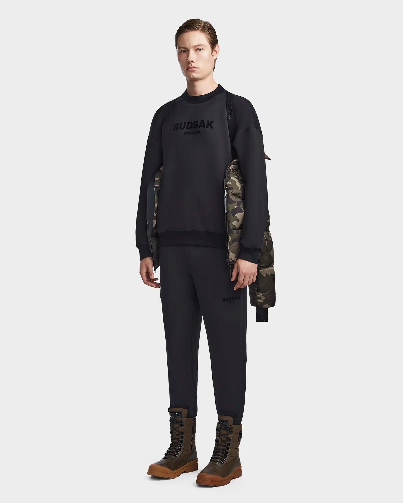 A person faces forward wearing the Rudsak RONNY M - M123512 Camouflage jacket, styled over a black sweatshirt and joggers with RUDSAK on the front and brown lace-up boots—showcasing sleek contemporary mens outerwear.