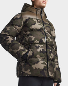 Wearing the RONNY M - M123512 Camouflage men’s hooded down puffer by Rudsak, featuring a green, brown, and black camouflage print and zippered pockets, this stylish outerwear is weather resistant and shown cropped from the nose down.
