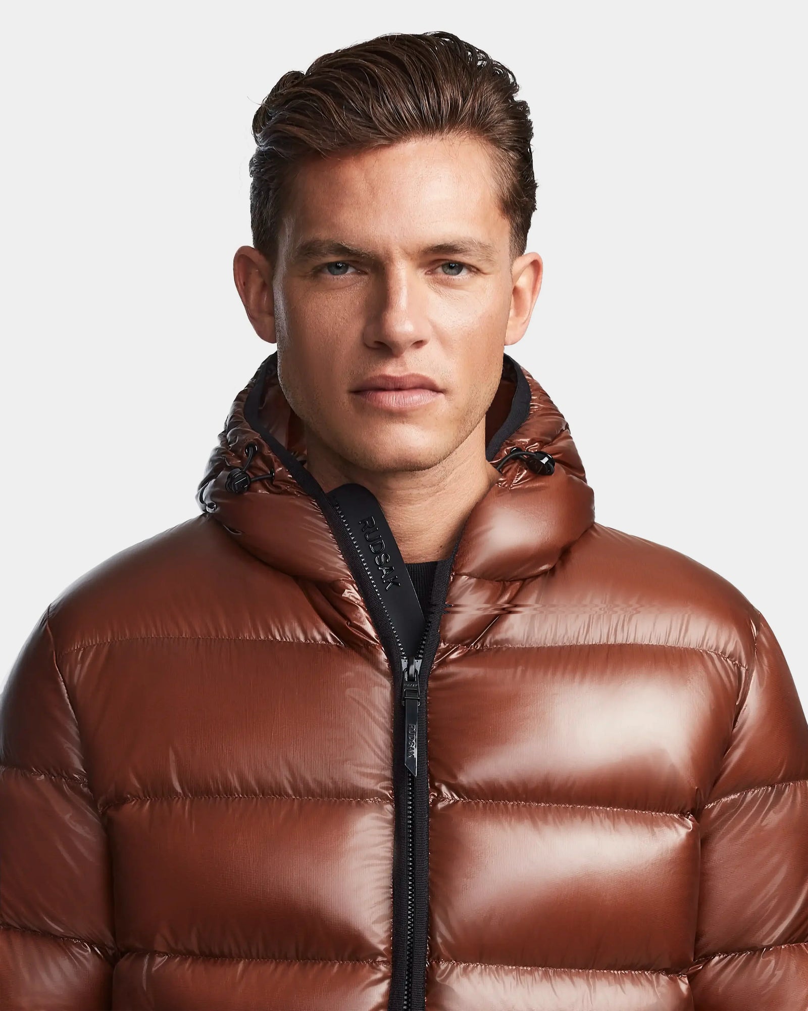 A man with short brown hair wears the Rudsak DEMIAN S - M123508 Cohiba, a shiny, water- and wind-resistant hooded down puffer jacket. It’s zipped up as he faces forward with a neutral expression against a plain light background.