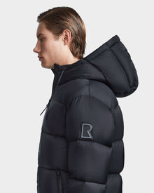 A young man with straight, light brown hair wears the Rudsak DANIEL M - M123510 BLACK mens down puffer with a large R patch on the upper sleeve, standing sideways against a plain white background.